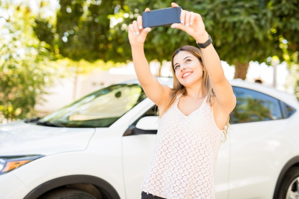 Woman in a white lace top taking a selfie with a smartphone outdoors beside a white car, smiling at the camera.