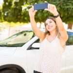 Woman in a white lace top taking a selfie with a smartphone outdoors beside a white car, smiling at the camera.