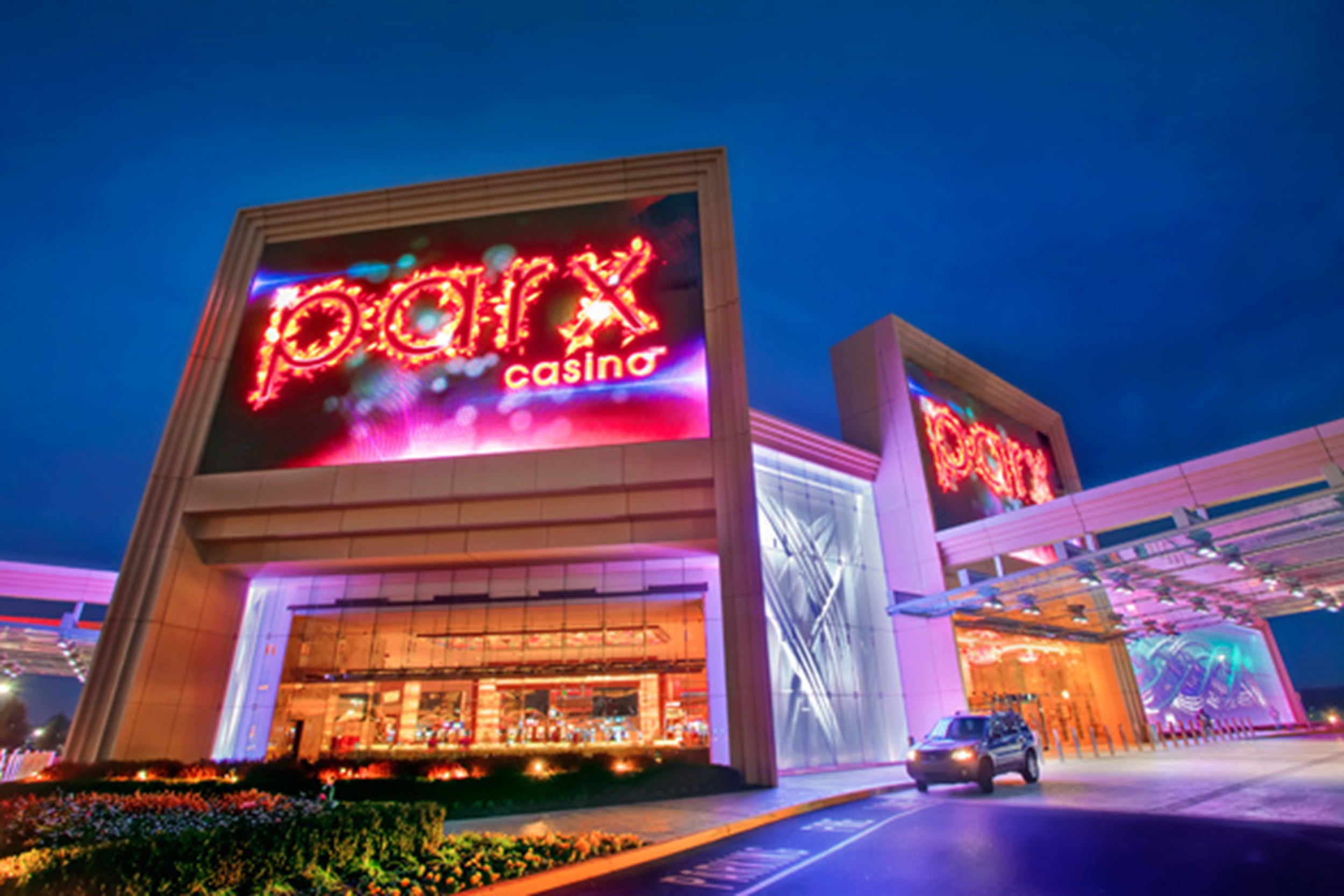 Exterior of a modern casino at night with red neon signage on a tall building and a car in the driveway.