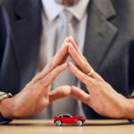Red toy car on a desk with two suited professionals in the background, their hands pressed together in a prayer-like gesture.