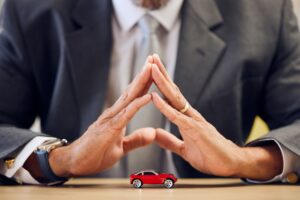 Red toy car on a desk with two suited professionals in the background, their hands pressed together in a prayer-like gesture.
