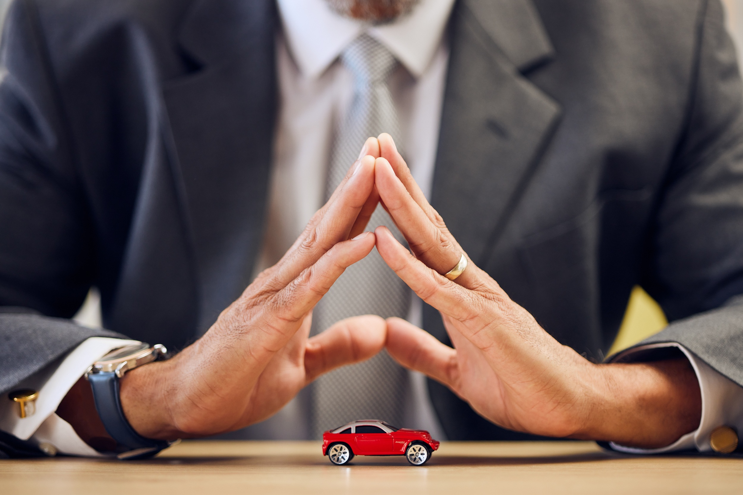 Red toy car on a desk with two suited professionals in the background, their hands pressed together in a prayer-like gesture.
