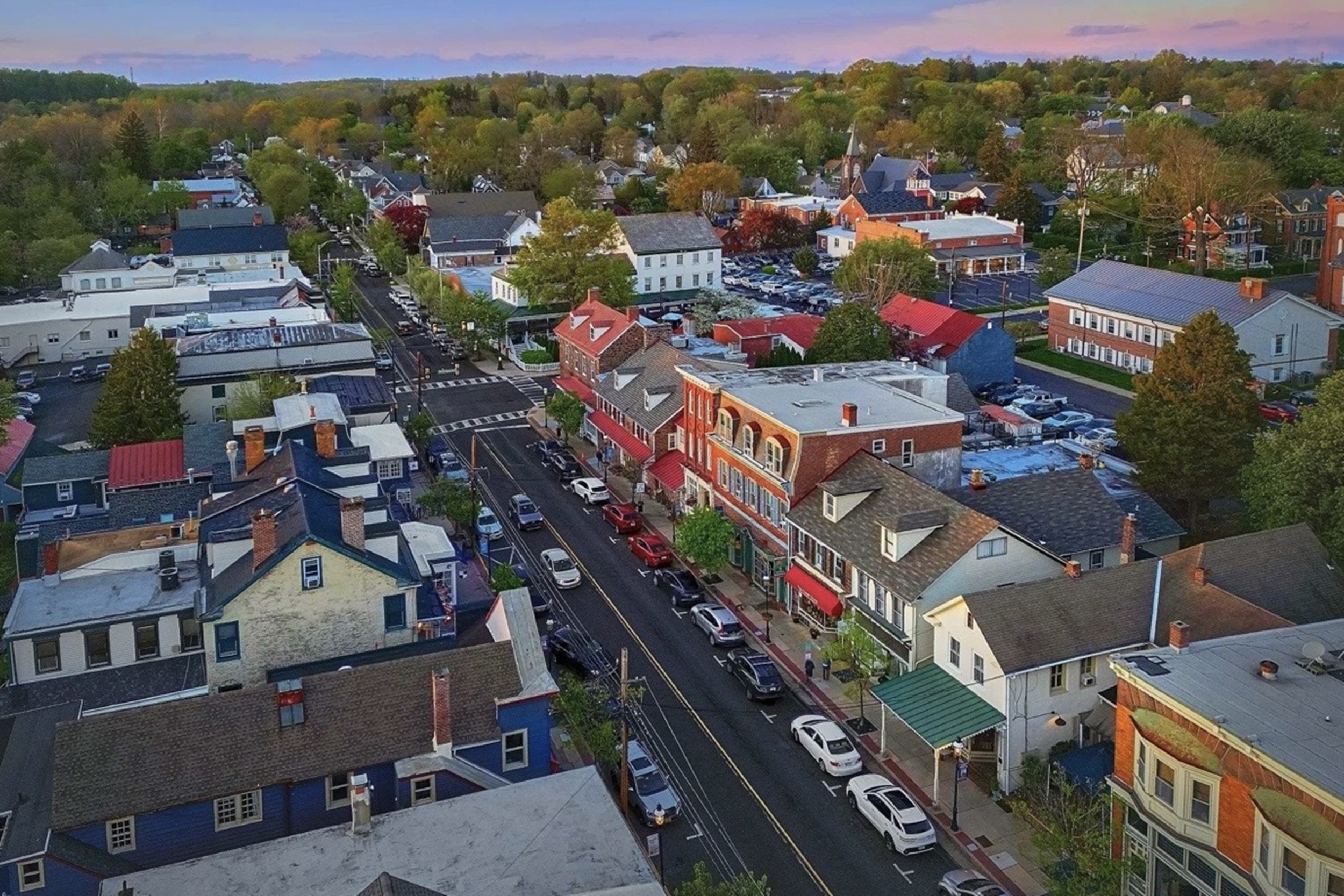 Aerial view of a small town with colorful storefronts along a main street, lined by trees and parked cars at sunset.