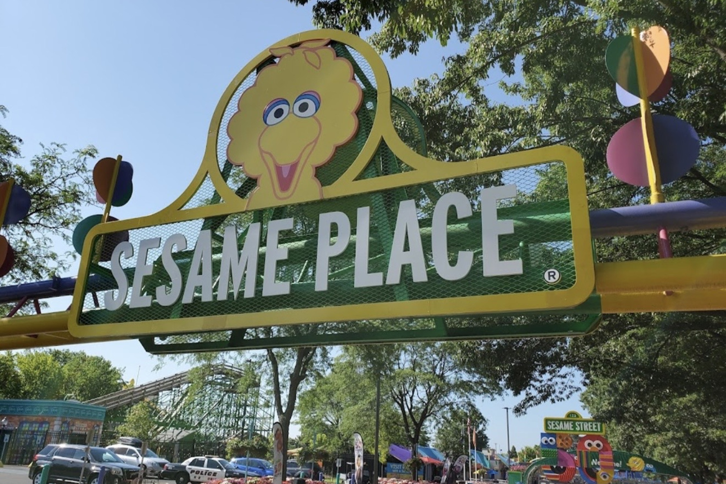 Entrance sign at Sesame Place featuring a large yellow cartoon character face and bold white letters on green background, with colorful balls and trees in the scene.
